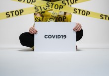 Osoba siedzi i trzyma kartkę z napisem Covid19. Przed nią żółte taśmy z napisem Stop.