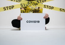 Na zdjęciu siedząca kobieta trzymające kartkę z napisem: 'covid19'. Dookoła taśmy z napisem: 'stop' - powiększ