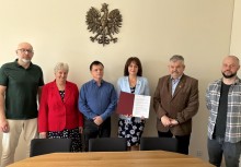 Na zdjęciu dwie kobiety i czterech mężczyzn. Burmistrz Gminy Żukowo trzyma podpisany dokument. Na ścianie zawieszone jest godło Polski. 