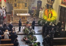 Na zdjęciu w centralnym miejscu trumna z kwiatami i zdjęciem w ramce. Wokół stojący ludzie.