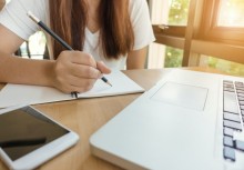 Kobieta notująca coś w notatniku, przed nią telefon i komputer - powiększ