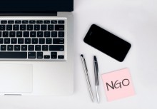 Na zdjęciu laptop, obok dwa długopisy, smartfon oraz różowa kartka na której znajduje się napis NGO