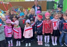 Zbliżenie na grupę dzieci z przedszkola w Miszewie – najmłodsi ubrani w kurtki i ozdobieni biało-czerwonymi szarfami oraz kokardami, uczestniczą w obchodach Święta Konstytucji 3 Maja na placu zabaw - powiększ