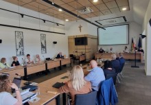 Sesja plenarna w sali obrad z udziałem kilkunastu osób siedzących przy stołach ustawionych w podkowę; na ścianie widoczny krzyż, ekran z prezentacją, zestaw flag oraz elementy nagłośnienia i oświetlenia. - powiększ