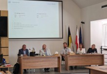 „Panel dyskusyjny z udziałem pięciu prelegentów podczas oficjalnego spotkania w sali konferencyjnej, z widoczną projekcją prezentacji, zestawem flag (w tym RP i UE) oraz elementami wystroju charakterystycznymi dla instytucji publicznej.”
