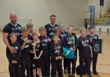 Drużyna młodych sportowców z trenerami po zawodach – wszyscy w czarnych strojach z medalami, pucharem i sportowym banerem.








 - powiększ