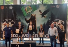 Ceremonia wręczenia medali na tle plakatu z niedźwiedziem – troje młodych zawodników na podium, w otoczeniu organizatorów. - powiększ