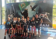 Grupa młodych zapaśników z trenerem po zawodach, pozująca na tle baneru z niedźwiedziem i orłem – wszyscy w strojach sportowych z medalami. - powiększ