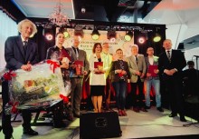 Grupa osób pozuje na scenie z wyróżnieniami i upominkami podczas uroczystej gali. Na środku stoi kobieta w białej marynarce, obok niej laureaci trzymający dyplomy i statuetki. W tle widoczny baner wydarzenia i kolorowe oświetlenie sceniczne - powiększ