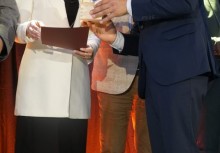 Zbliżenie na moment wręczenia dokumentu – kobieta w białej marynarce i mężczyzna w granatowym garniturze uczestniczą w oficjalnej ceremonii na tle czerwonej kotary.








 - powiększ