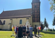 Grupa osób pozuje do zdjęcia przed kościołem z drewnianą wieżą i figurą Matki Bożej. Obok znajdują się flagi narodowe i kościelne, a pogoda jest słoneczna. Wśród uczestników widoczny jest duchowny, osoby w strojach oficjalnych i wojskowych. - powiększ