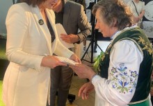 Kobieta w białej marynarce wręcza dyplom starszej uczestniczce w tradycyjnym kaszubskim stroju ludowym. Obie uśmiechają się, a w tle widoczni są inni goście i elementy dekoracji regionalnej. - powiększ
