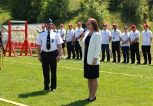 Kobieta w białej marynarce przemawia do mikrofonu na boisku sportowym, obok niej stoi strażak w galowym mundurze. W tle ustawieni w szeregu uczestnicy wydarzenia w białych koszulkach i czerwonych czapkach. Pogoda słoneczna, sceneria zielona - powiększ