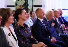 Uczestnicy wydarzenia siedzą w pierwszym rzędzie, skupieni i uśmiechnięci. Wśród nich kobieta w białej marynarce oraz mężczyźni w garniturach. W tle widoczna publiczność i elementy dekoracji wnętrza. - powiększ