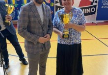  Wręczenie pucharu – uśmiechnięta kobieta trzyma trofeum podczas wydarzenia w hali sportowej. Obok niej stoi mężczyzna w marynarce. W tle baner UKS Banino i plansza „Sportowe podsumowanie roku Gminy Żukowo”. - powiększ