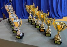  Rząd pucharów ustawionych na stole – złote i srebrne trofea przygotowane na uroczystość sportową lub edukacyjną w hali. W tle granatowa kurtyna sceniczna. - powiększ