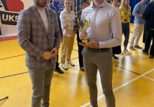  Wręczenie pucharu – mężczyzna odbiera trofeum podczas uroczystości w hali sportowej. W tle uczestnicy wydarzenia i elementy baneru UKS Banino. - powiększ