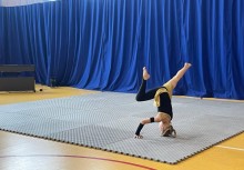  Pokaz gimnastyczny – dziewczynka w stroju sportowym wykonuje efektowną figurę na macie podczas występu w hali sportowej. W tle granatowa kurtyna sceniczna. - powiększ