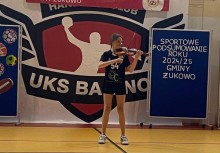 Występ artystyczny podczas wydarzenia „Sportowe podsumowanie roku 2024/25 Gminy Żukowo” – dziewczynka w stroju sportowym gra na skrzypcach na tle baneru UKS Banino. - powiększ