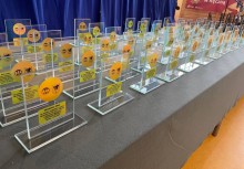Statuetki przygotowane na uroczystość – przezroczyste trofea z żółtym emblematem, ustawione w rzędach na stole.