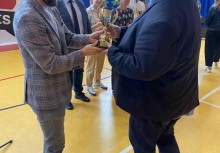 Wręczenie nagrody – mężczyzna w jasnej marynarce przekazuje puchar drugiemu uczestnikowi wydarzenia. Uroczystość odbywa się w hali sportowej, w tle obecni są inni uczestnicy i baner UKS Banino.
 - powiększ