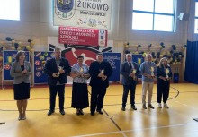 grupa siedmiu osób pozuje z nagrodami na tle baneru „Gmina Żukowo” i „Gminny Konkurs Wiedzy o Młodzieży i Środowisku”. Uroczystość odbywa się w hali sportowej, dekoracje wskazują na podsumowanie sportowego roku szkolnego. - powiększ