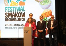 Kobieta przy mównicy z napisem 'amberexpo', za nią stoi trzech mężczyzn. W tle widać duży baner z napisem 'Festiwal Smaków Regionalnych'.  - powiększ