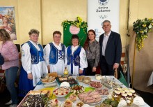 Zdjęcie grupowe przy stole z potrawami Wielkanocnymi. W tle plakat z logo Gminy Żukowo. - powiększ