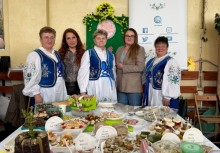 Zdjęcie grupowe przy stole z potrawami Wielkanocnymi. W tle plakat z logo Gminy Żukowo. - powiększ