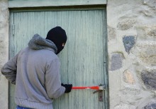 Na zdjęciu człowiek w kominiarce z łomem stara się otworzyć drzwi. - powiększ