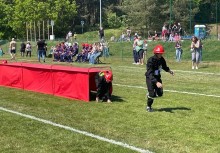 Dzieci w strażackich strojach pokonują przeszkody podczas zawodów sportowo-pożarniczych na boisku. - powiększ