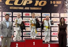 Trzy młode z zawodniczki w strojach do judo stoją na podium z medalami i pucharami. Po bokach dwoje dorosłych. W tle duży baner z logo 'Same Judo' i logotypami sponsorów.  - powiększ