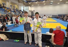 Na pierwszym planie dwóch chłopców z dyplomami. W tle mata, na której widać zawodników. Po lewej stronie widownia   - powiększ