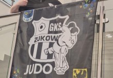 Na pierwszym planie flaga GKS Żukowo Judo zawieszona na barierce. Za nią stoi osoba pokazując kciuk w górę. - powiększ