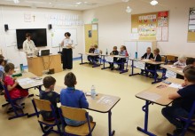 Dzieci siedzą przy stolikach podczas konkursu. Dwie kobiety stoją przy biurku