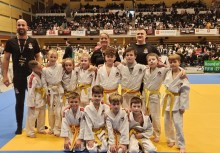 Grupa dzieci w strojach do judo oraz troje dorosłych. W tle widać trybuny z publicznością.  - powiększ