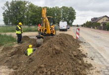 Dwóch pracowników w kaskach i kamizelkach odblaskowych pracuje przy wykopie ziemnym przy drodze. W tle widać budynki, drzewa oraz zaparkowane samochody. 