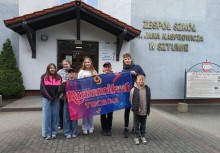 Siedem osób trzymających flagę z napisem Rummikub. W tle budynek Zespołu Szkół w Sztumie - powiększ
