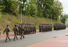 Liczna grupa młodzieży ubrana w mundury i berety stoi w dwuszeregu. Przed nimi maszerują trzy umundurowane osoby. W tle widoczne są drzewa rosnące na wzgórzu. - powiększ