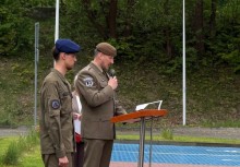 Mężczyzna w mundurze i berecie przemawia do mikrofonu, stojąc przy mównicy. Obok niego stoi młody chłopak w mundurze i granatowym berecie. W tle widoczne są drzewa. 