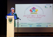 Kobieta przy mównicy z herbem Gminy Żukowo. W tle widać wyświetloną prezentację z napisem 'ZROZUMIEĆ AUTYZM aby skutecznie pomagać - X jubileusz'. 