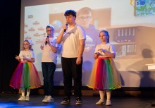 Dwóch chłopaków śpiewa do mikrofonu, a obok nich tańczą dwie dziewczynki w tęczowych spódniczkach. W tle wyświetlany jest filmik nagrany przez SOSW w Żukowie.  - powiększ