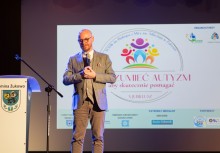 Mężczyzna przemawia do mikrofonu, obok niego stoi mównica z herbem Gminy Żukowo. W tle widać wyświetloną prezentację z napisem 'ZROZUMIEĆ AUTYZM aby skutecznie pomagać - X jubileusz'.  - powiększ