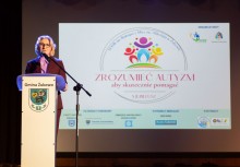 Kobieta przy mównicy z herbem Gminy Żukowo. W tle widać wyświetloną prezentację z napisem 'ZROZUMIEĆ AUTYZM - aby skutecznie pomagać - X jubileusz'.  - powiększ